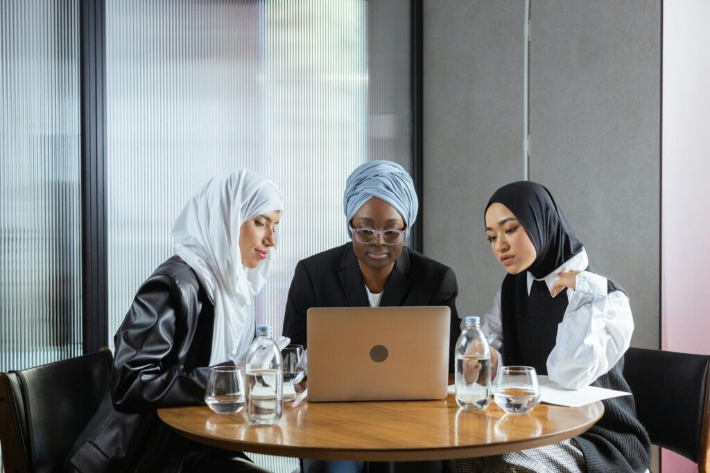 Three diverse women in hijabs and turbans collaborate indoors at a laptop for a professional meeting.